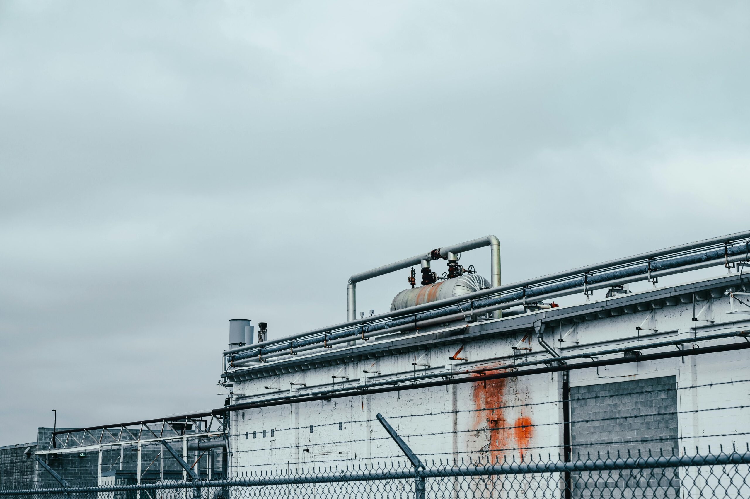 Exterior view of an industrial facility with metal piping and a fenced perimeter under an overcast sky.
